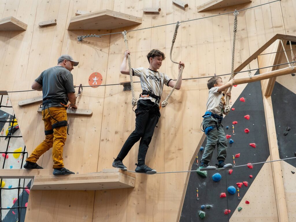 Barn og voksen balanserer på høydeløype innendørs hos Høyt Under Taket med sikringsutstyr og klatrevegg i bakgrunnen
